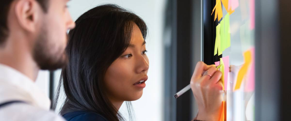 Focused asian business woman mentor coach leader writing idea or task on post it sticky notes on glass wall.