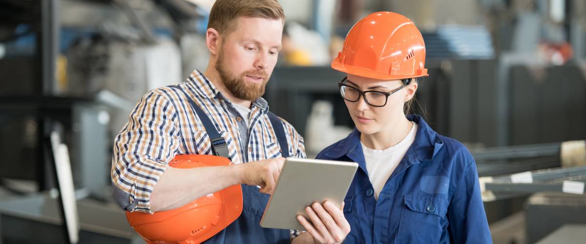 Young female engineer in uniform and helmet looking at tablet display while listening to colleague explanation of data