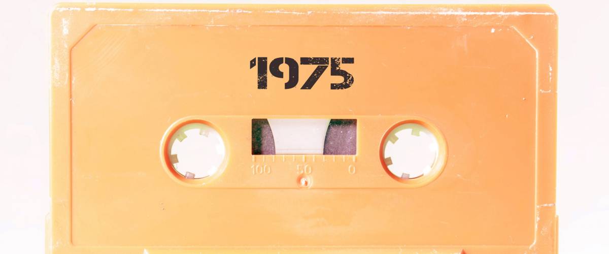 A vintage cassette tape from the 1980s era (obsolete music technology) with the text 1975 printed over it, stencil font. Color: cream, sand. White background.