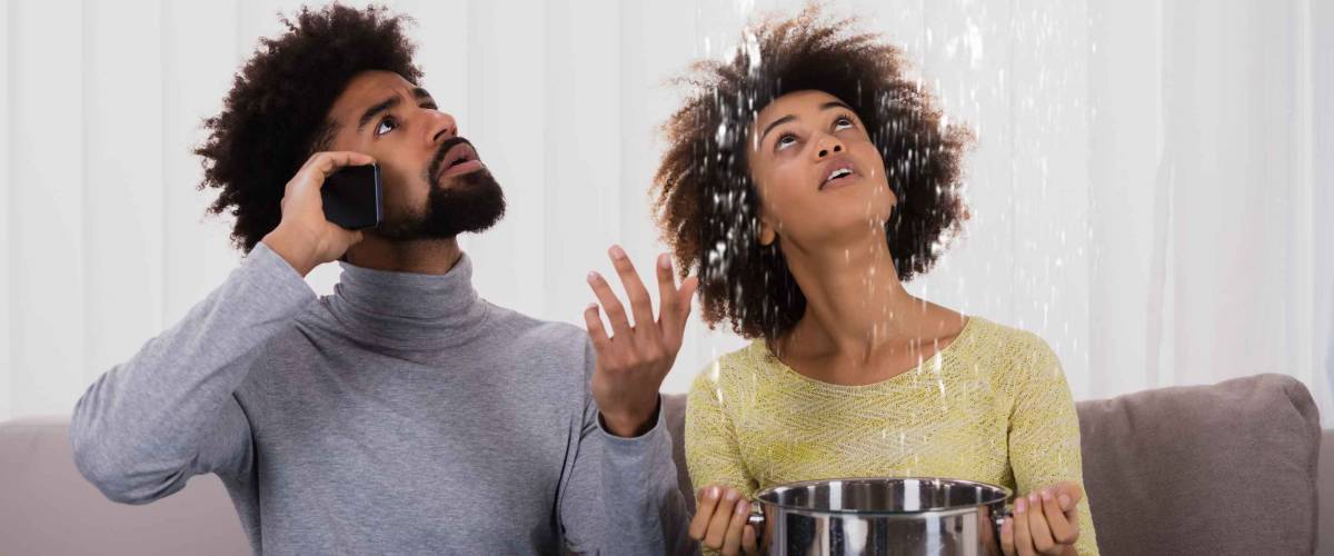 Young Man Calling Plumber While Woman Using Utensil For Collecting Water Leaking From Ceiling