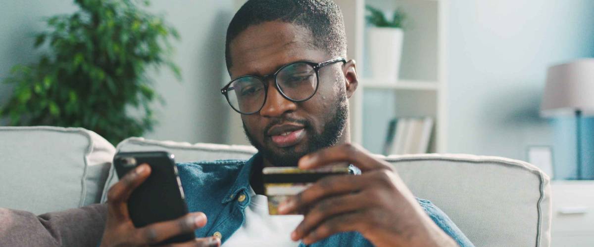 African young man in glasses shopping online with credit card using smart phone at home. Indoor.
