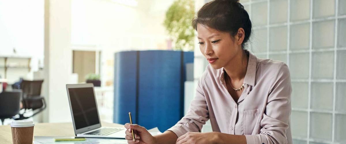 Young Asian designer sitting alone at a desk in an office looking over building designs and working on a laptop