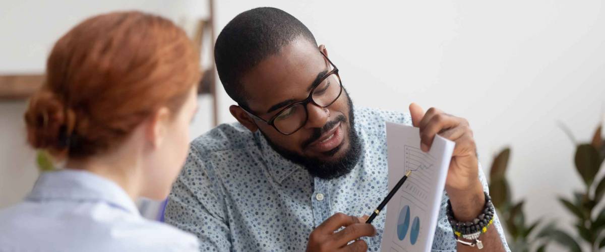 Male black coworker showing document with project results talking to caucasian female colleague.