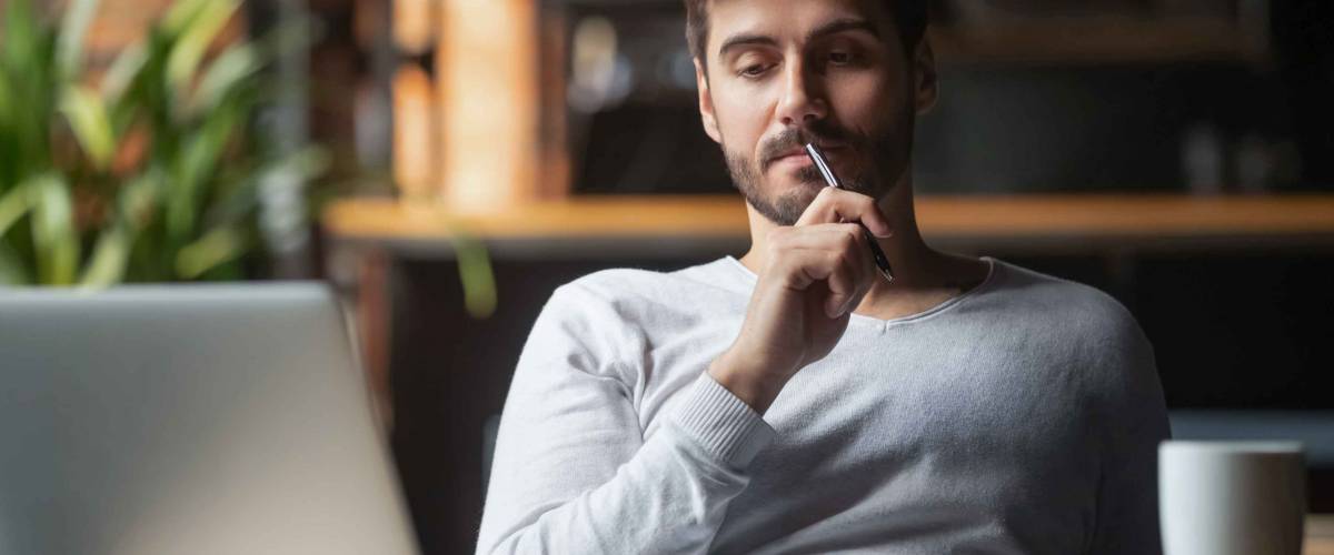 Pensive bearded man sitting at table drink coffee work at laptop thinking of problem solution