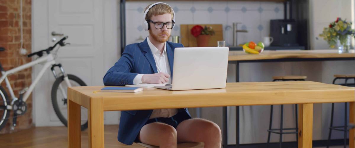 Young businessman wearing no pants and listening to headphones sitting at desk working remotely