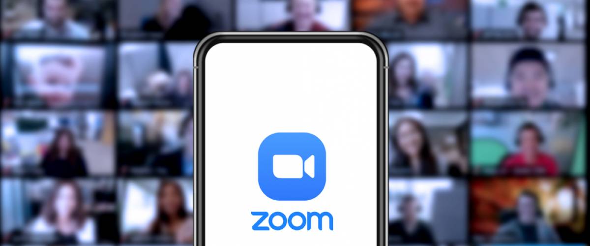 The Zoom logo on a cell phone screen in front of a bigger screen showing people in videoconference call.
