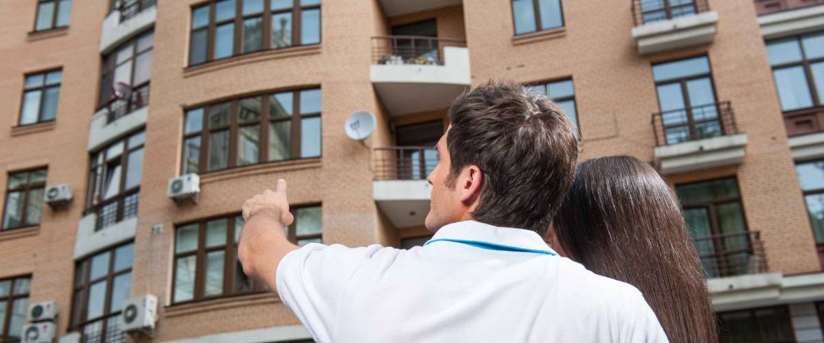 couple standing with their backs towards camera. man pointing to apartment window for brunette woman