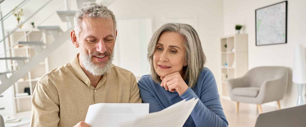 Middle aged senior old couple holding documents reading paper bills paying bank loan online