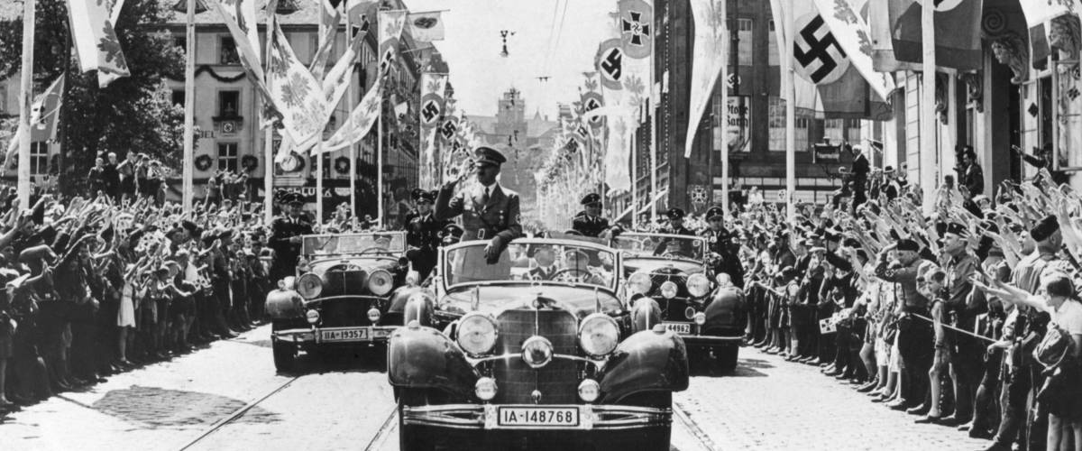 Adolf Hitler waving to crowds from his car at the head of a parade. The streets are decorated with various swastika banners. Ca. 1934-38. Location is unidentified.