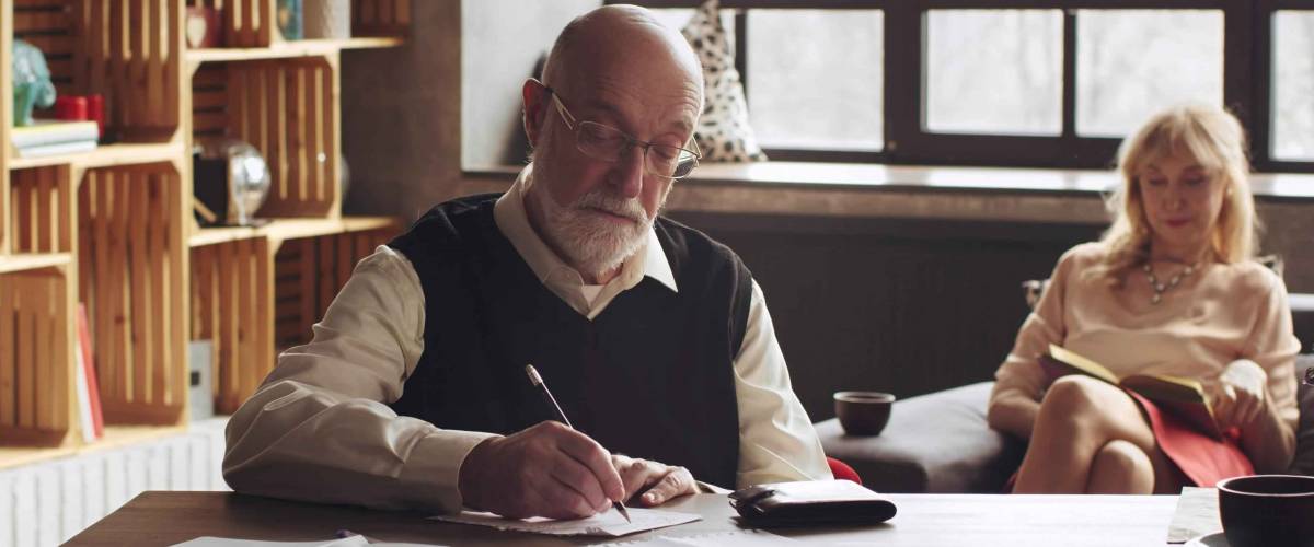 Senior man writing a letter sitting at desk in living room with aged wife reading book on sofa in background.