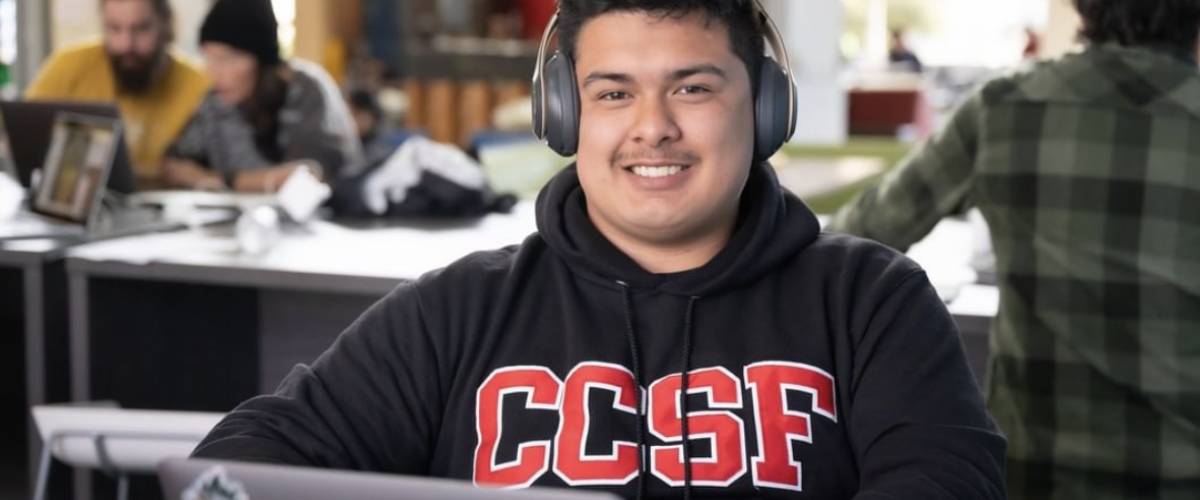 Young man wearing CCSF sweatshirt, sits with headphones on a laptop looking at camera