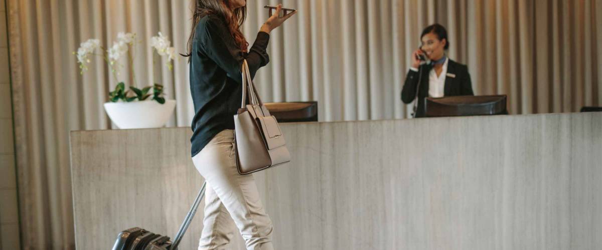 Business woman walking in front of hotel reception with luggage and having a phone call. Female guest arriving in hotel with suitcase.