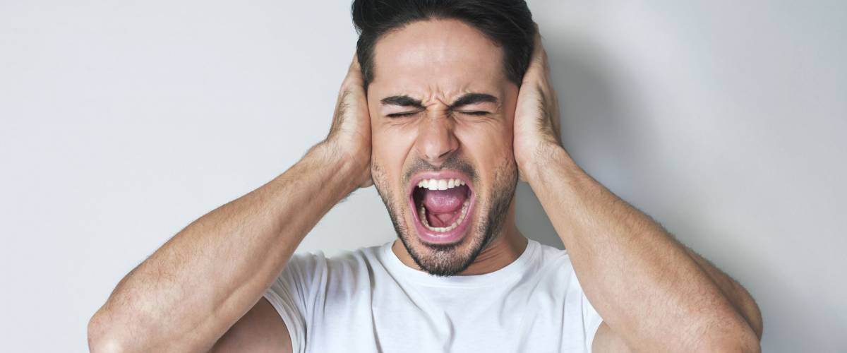 Stop that loud noise! Standing plain young man sad and depressed suffering sorrow and pain screaming desperate with hands on face against grey background. Sadness emotion concept