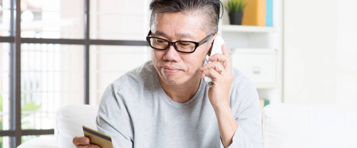 Asian mature man making order by phone, looking at credit card, and using laptop computer at home.