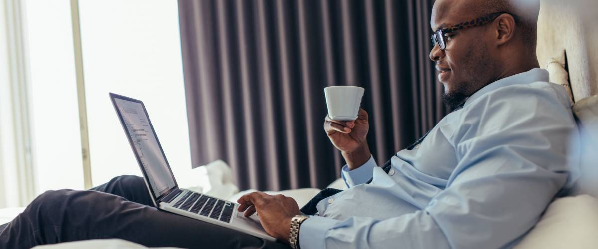 Man in formal clothes working on laptop while lying in bed. Businessman working on laptop computer while drinking coffee in his hotel room.