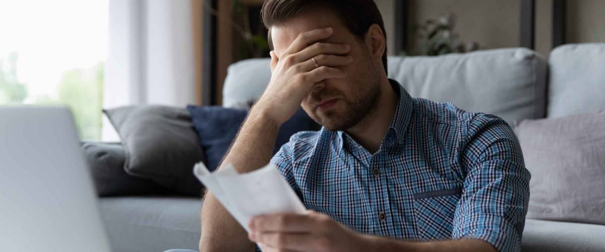 Frustrated desperate millennial man checking bills for payments, holding piece of paper