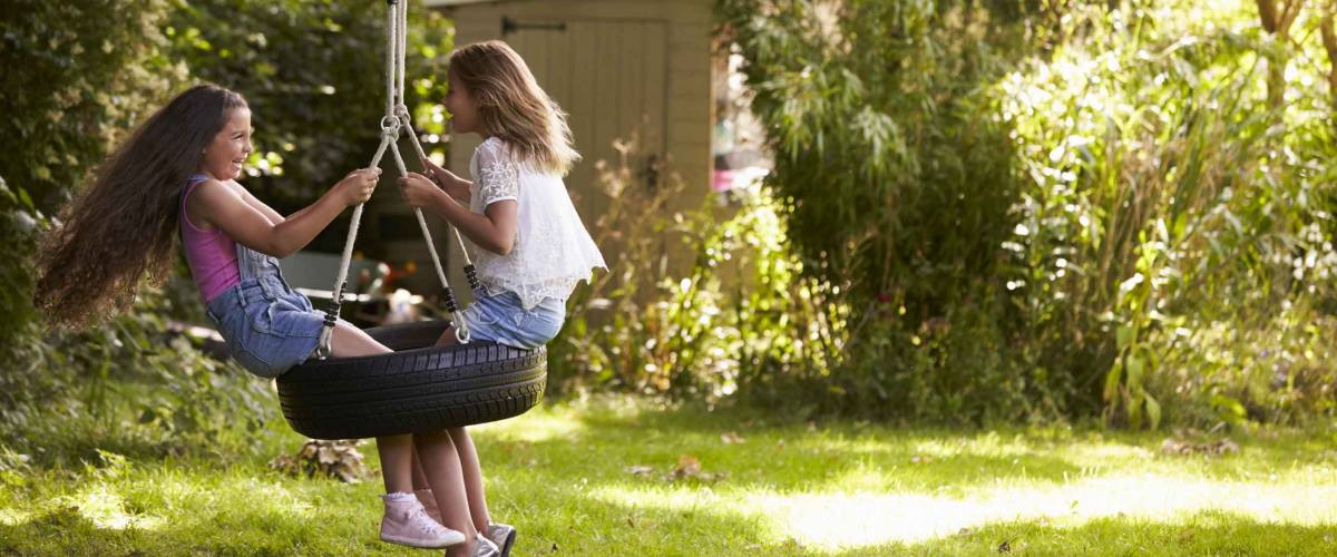 Two Girls Playing Together On Tire Swing In Garden