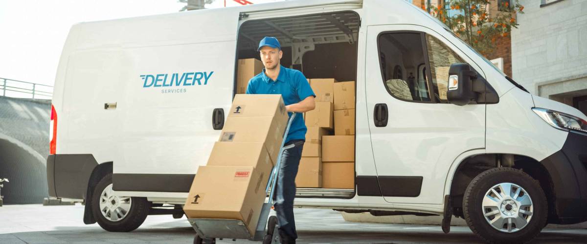 Delivery Man Pushes Hand Truck Trolley Full of Cardboard Boxes Hands Package to a Customer. Courier Delivers Parcel to Man in Business District.