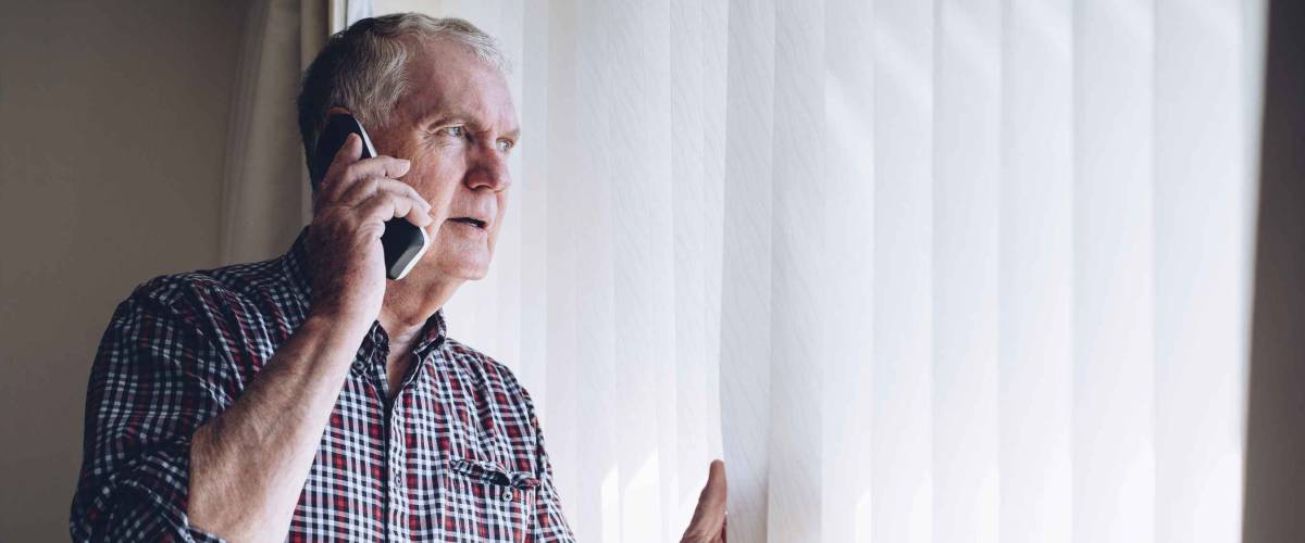 Senior man talking on the phone whilst looking out of his window. He has a worried look on his face.
