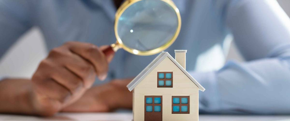Close-up Of A Businesswoman's Hand Holding Magnifying Glass Over House Model Over Desk