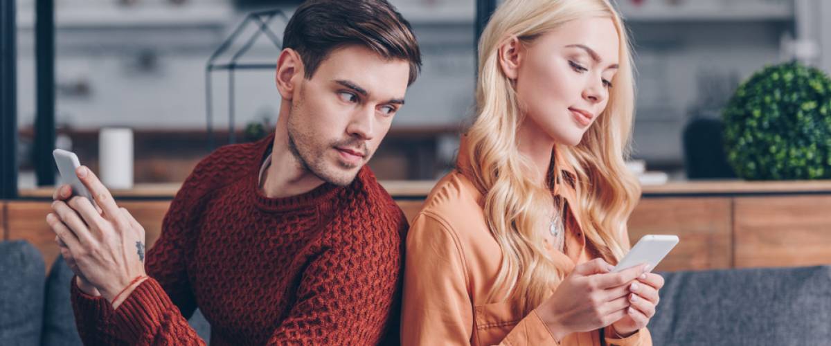 Young couple sitting back to back, man jealously looking at woman's phone
