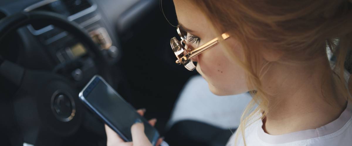 Blond teenager wearing sunglasses on her phone while driving.