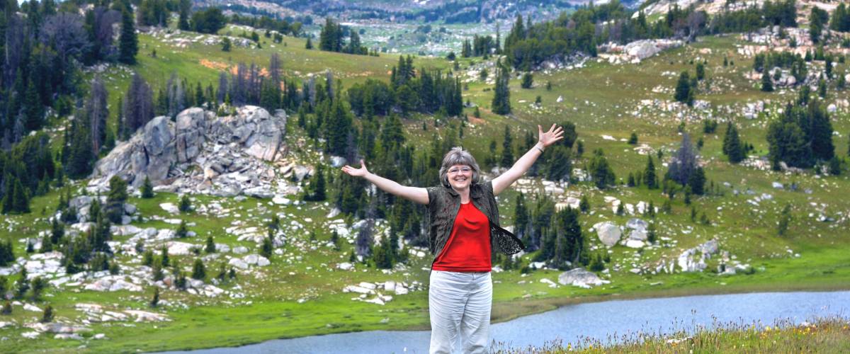 Woman spreads her arms wide to embrace the scenic beauty of Beartooth Pass in Wyoming. She has on a red shirt and khaki pants.