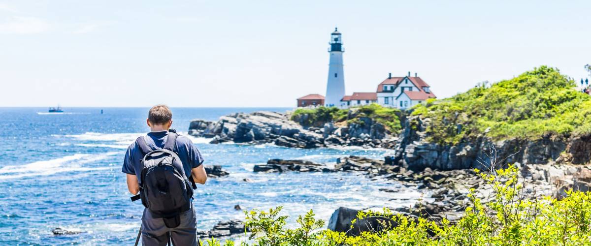 Back of man photographer taking pictures on tripod of Portland Head Lighthouse in Fort Williams park in Maine during summer day