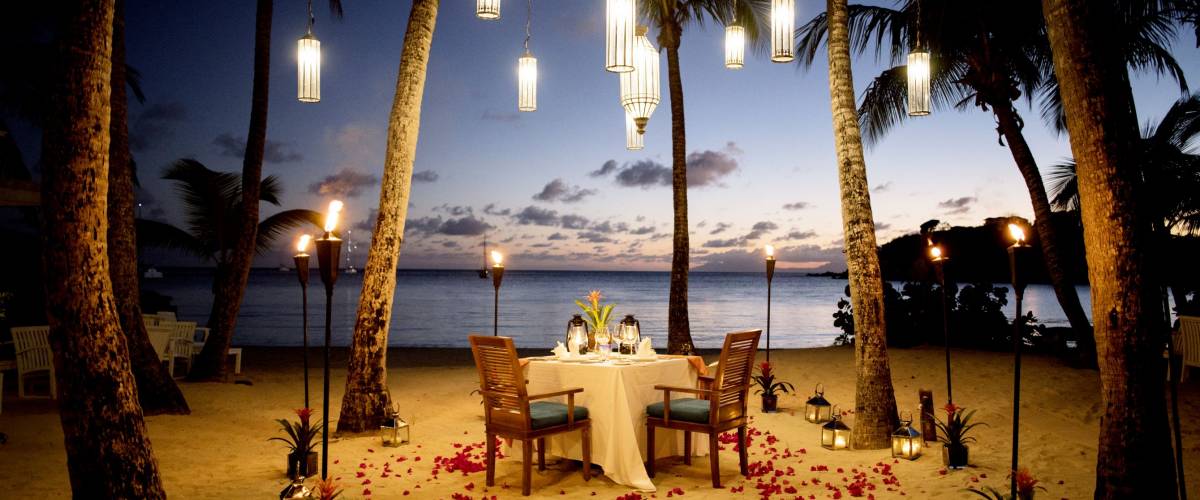 A Table Set up for a romantic meal on the beach with lanterns and chairs and flowers with palms and sky and sea in the background