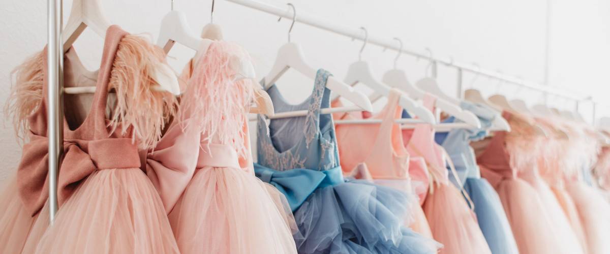 Beautiful dressy lush pink and blue dresses for girls on hangers at the background of white wall. Kids dresses with feathers for prom and holiday.