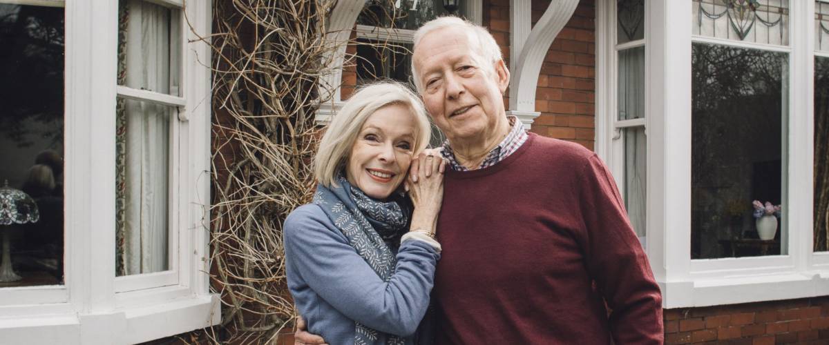 Senior couple are smiling for the camera while standing in front of their home.