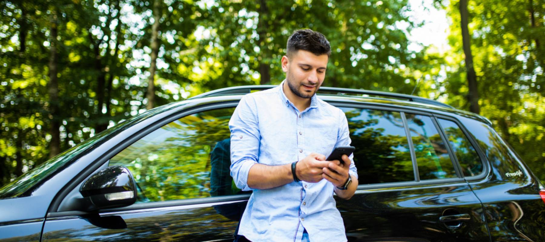 Young attractive smiling man with beard standing near his car and holding mobile phone