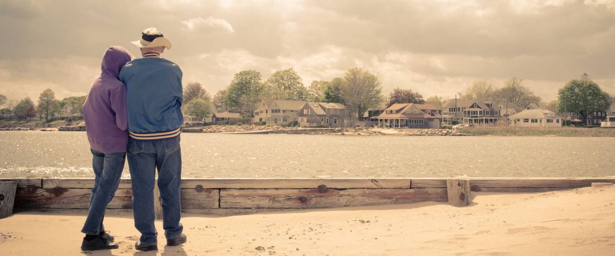 An older couple on the beach looking across the water at beautiful waterfront homes. Vintage effect applied.