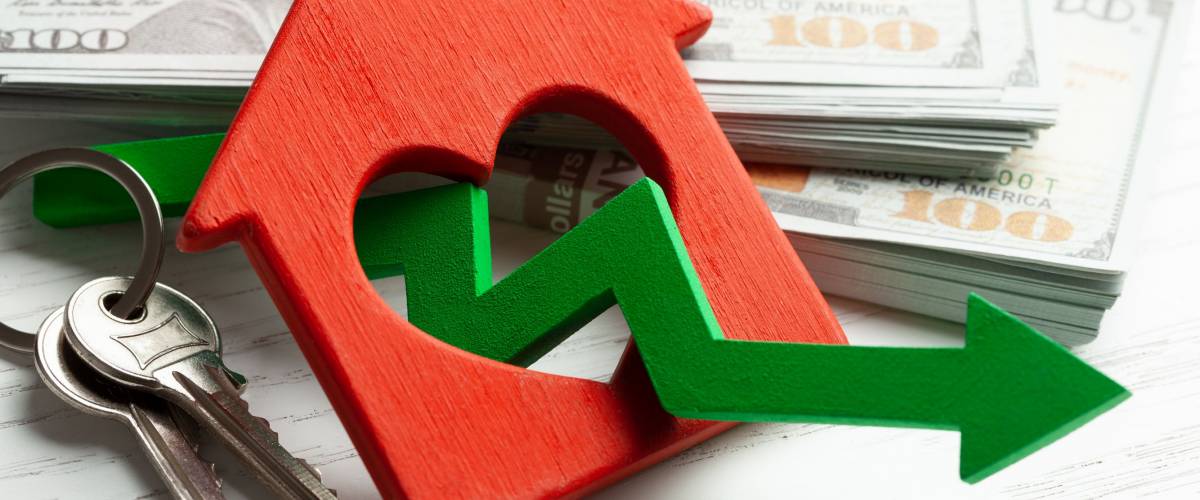 Arrow down and house with money on white wooden background. Falling market price of real estate.
