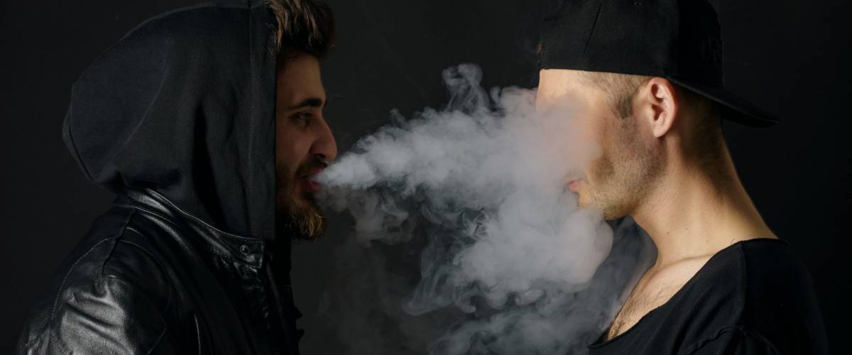 Bad habit, smoking in a public place. Two Young man blowing smoke to join it in one cloud at black studio background. Friends and vape addiction concept.