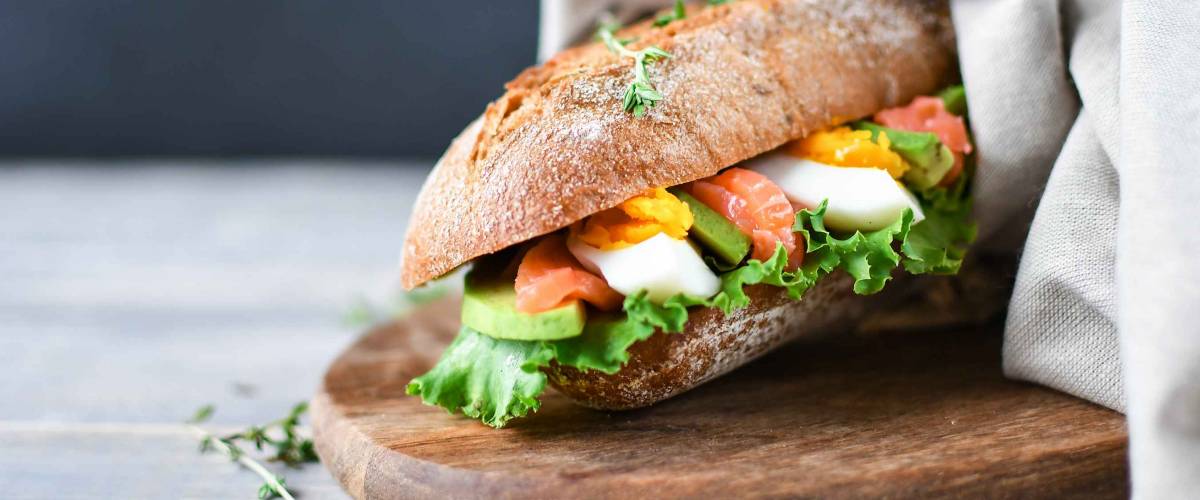 sandwich from a cereal baguette with avocado, salmon, egg and lettuce leaves on a wooden plate. wooden table and dark background, selective focus and copy space