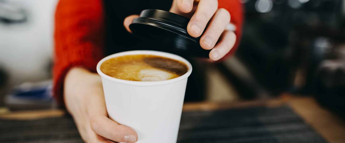 Barista in apron is holding in hands hot cappuccino in white takeaway paper cup. Coffee take away at cafe shop