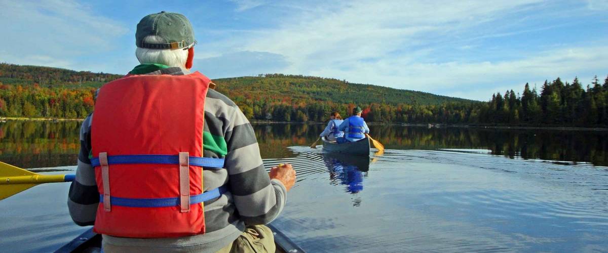 Senior man paddling a canoe with second canoe, calm waters and autumn colors in background.