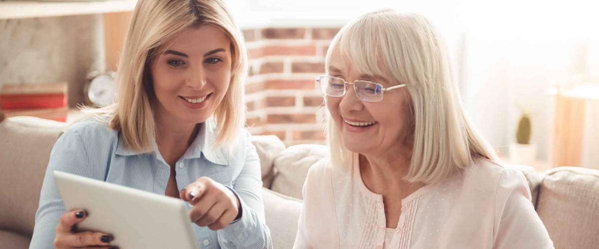 Beautiful mature mother and her adult daughter are drinking coffee, using a digital tablet and smiling while sitting on couch at home