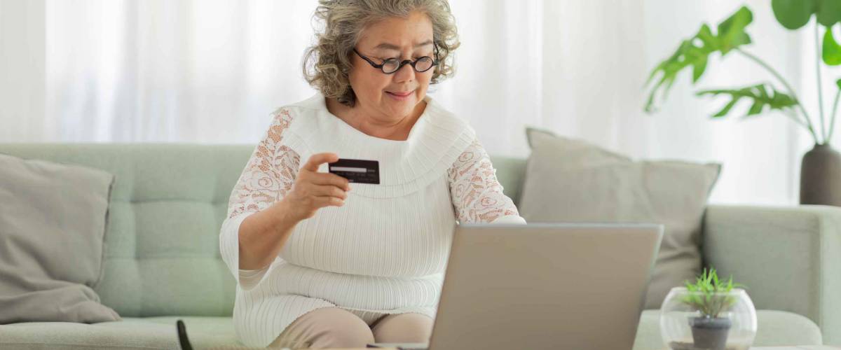 elderly asian woman with white hairs sitting on sofa using computer laptop and using credit card to shop online