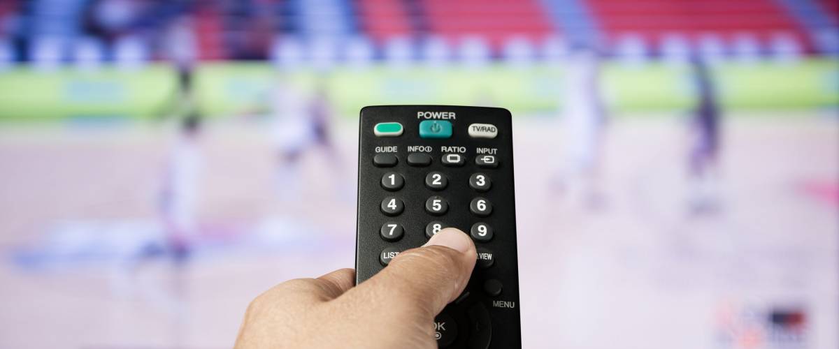 Hand of man holding remote control TV on sport channel program background
