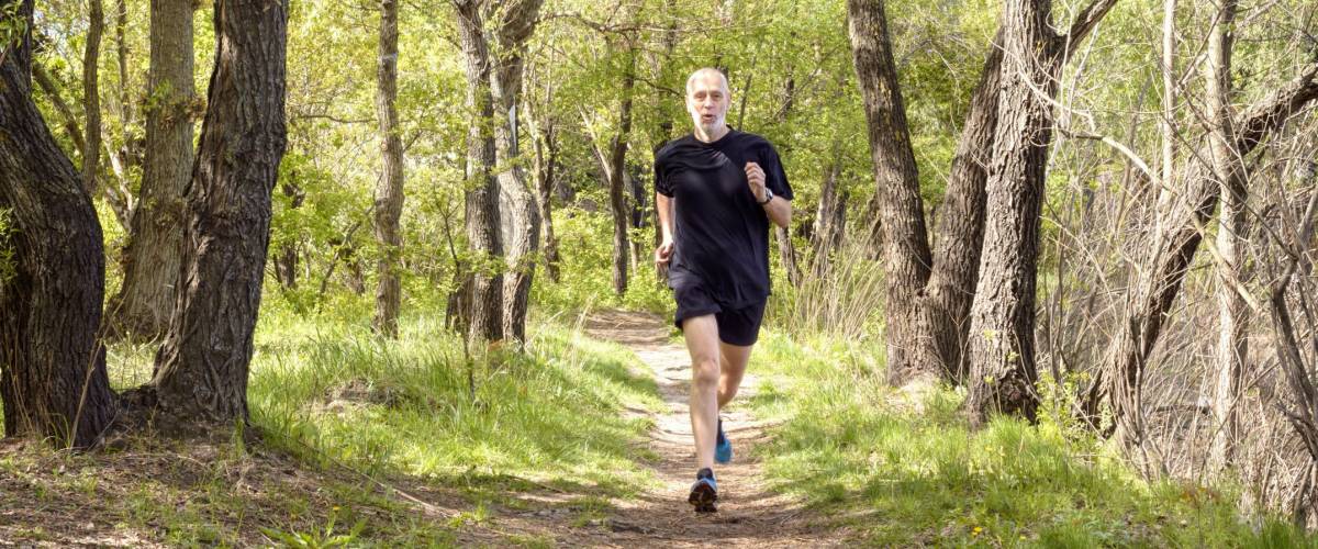 A senior man dressed in black is running in the forest during a warm spring day