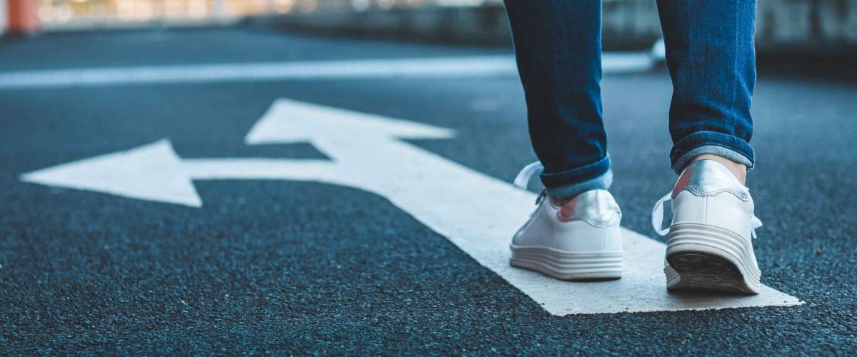 Make decision which way to go. Walking on directional sign on asphalt road. Female legs wearing jeans and white sneakers.