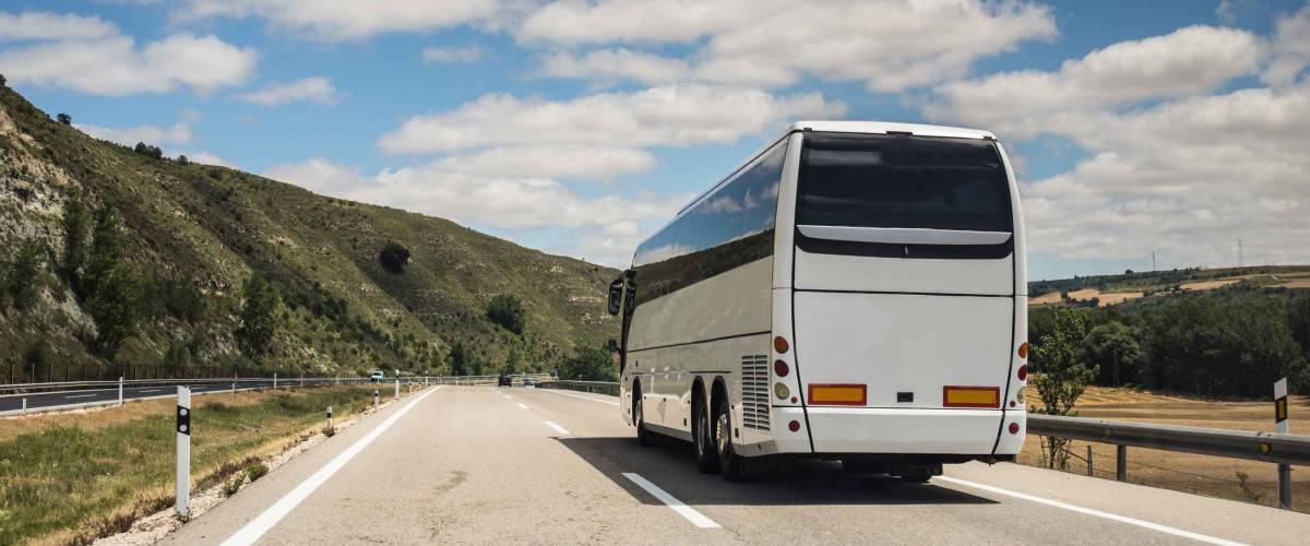 A white coach, or long haul bus for tourists drives through the open roads of Spain, Europe on a summer day. There are white clouds against a blue sky and countryside.