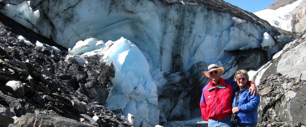Older couple stand in front of the Worthington Glacier in Alaska. The man is wearing a red jacket and the woman a blue.
