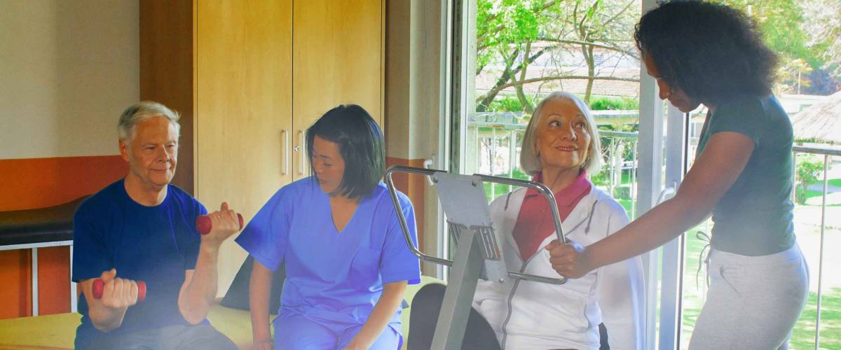 Rehabilitation concept. Elderly retired couple training in a gym supervised by expert african and asian female trainers.