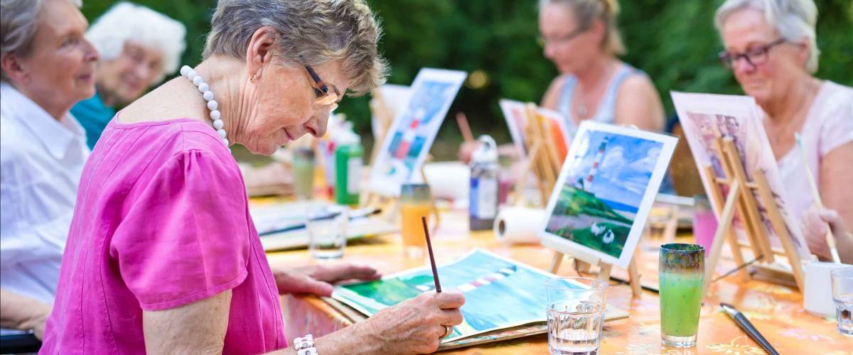 Stylish senior lady painting in art class with friends from her care home for the aged copying a painting with water colors on a canvas outdoors at a table in the garden.