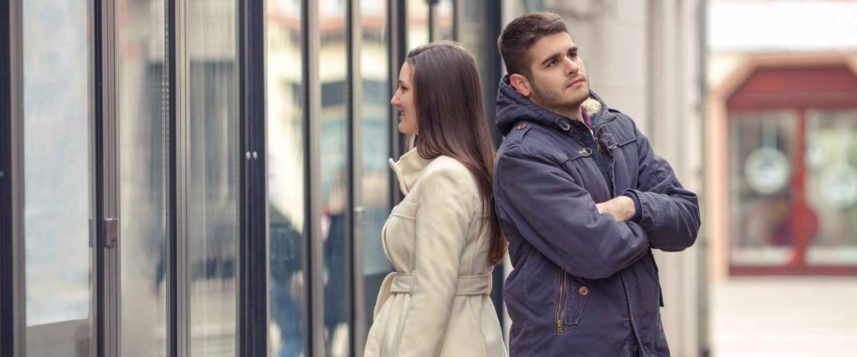 Young couple standing in front of the boutique. Girl looking at store window and bored young man standing behind her back with his arms crossed waiting for her.