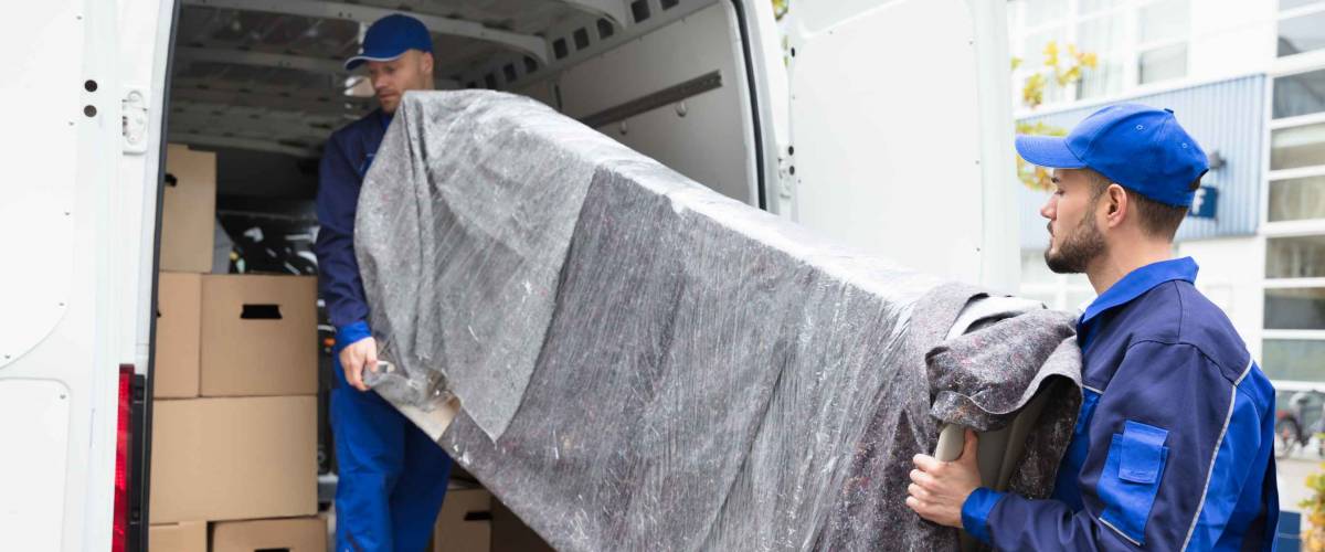 Two Young Delivery Men In Uniform Unloading Furniture From Vehicle