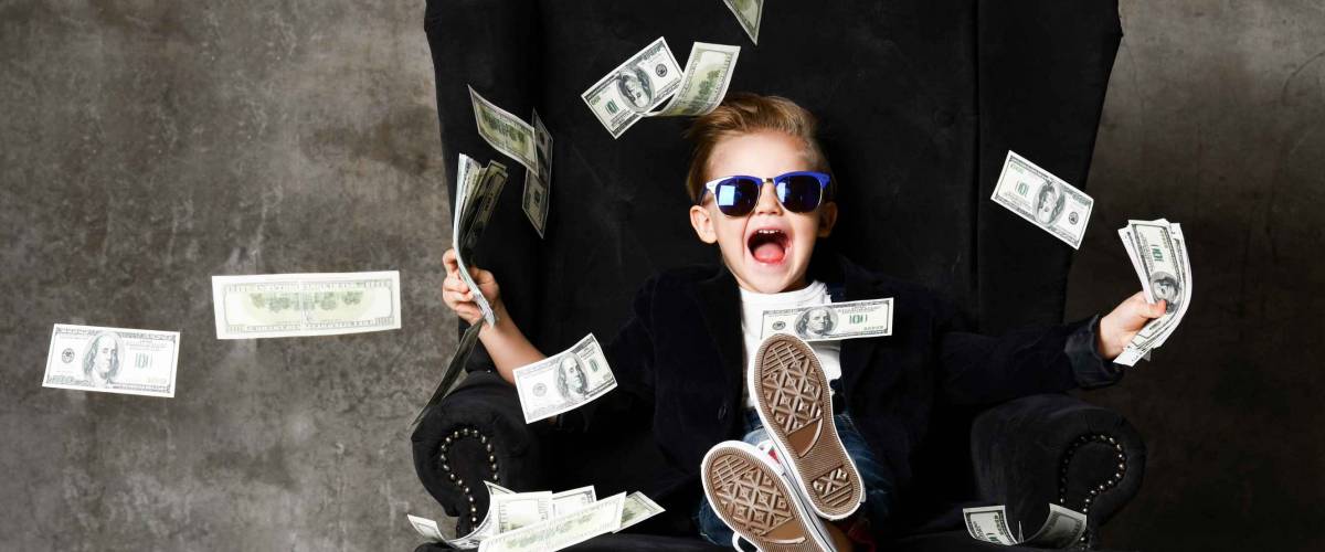 Happy shouting self-confident rich kid boy millionaire sitting with legs crossed in luxury armchair and throwing money dollars cash. Overspend.